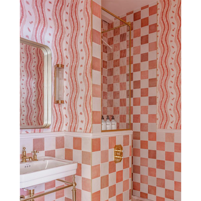A pink and white checkered tiled bathroom corner with wavy pink wallpaper, a white sink, gold fixtures, and a glass shower door. Our Ikat stripe wallpaper is sold by the metre online and from our Kensington based showroom.