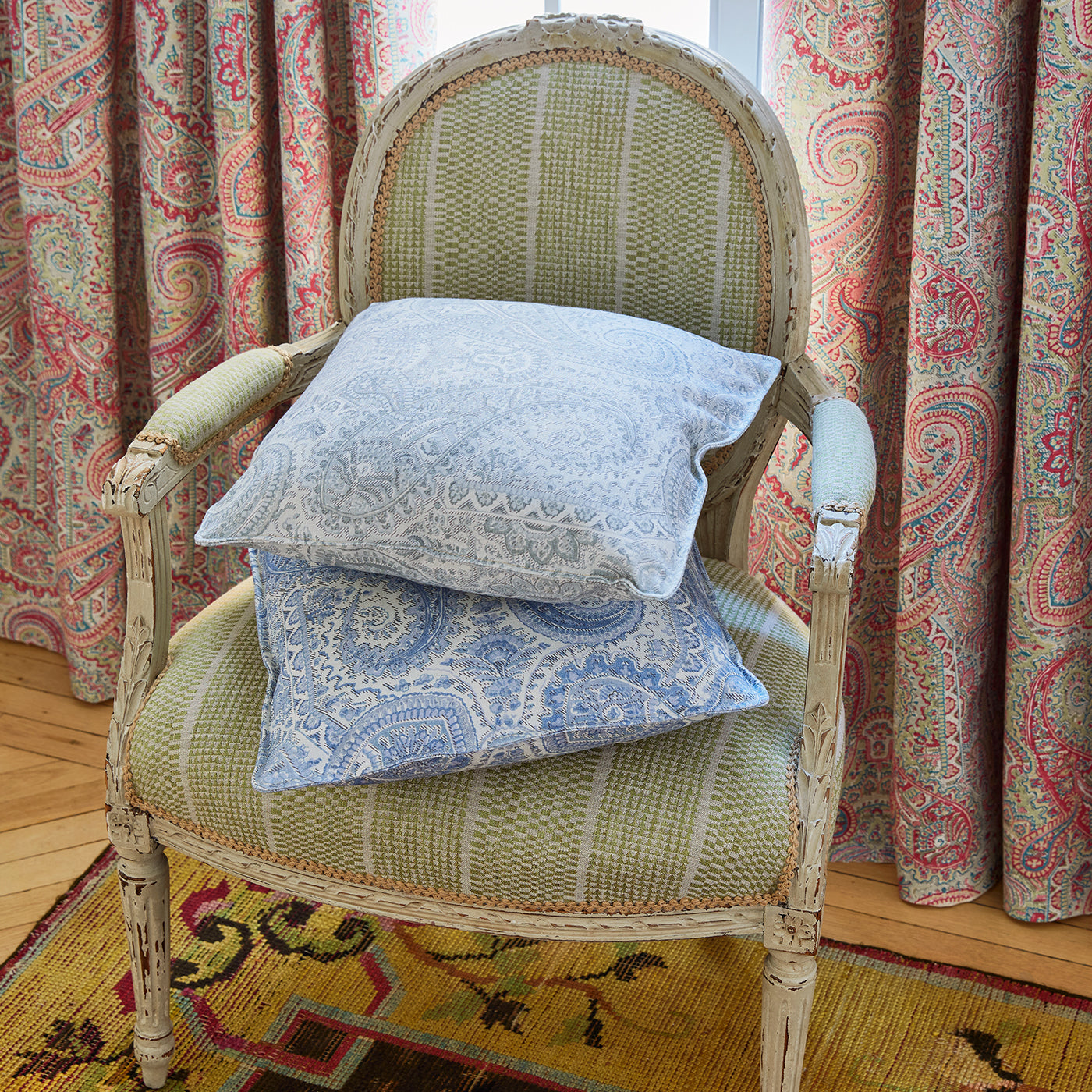 Two decorative cushions with paisley and patterns stacked on a distressed white wooden armchair upholstered in a green woven stripe fabric, Larkin in front of pink paisley patterned curtains.
