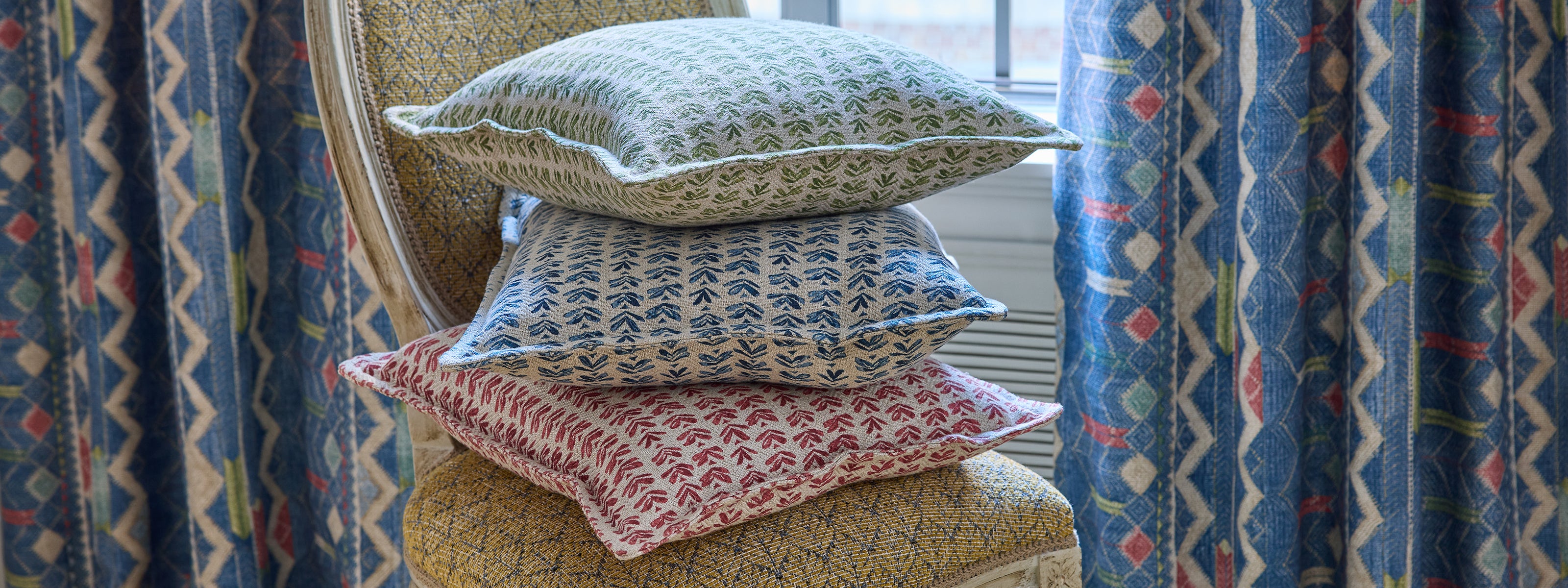 Three decorative cushions with a small-scale print in blue, red, and green stacked on an upholstered armchair in ochre. Luxury interior design fabrics sold by the metre online and our London showroom.