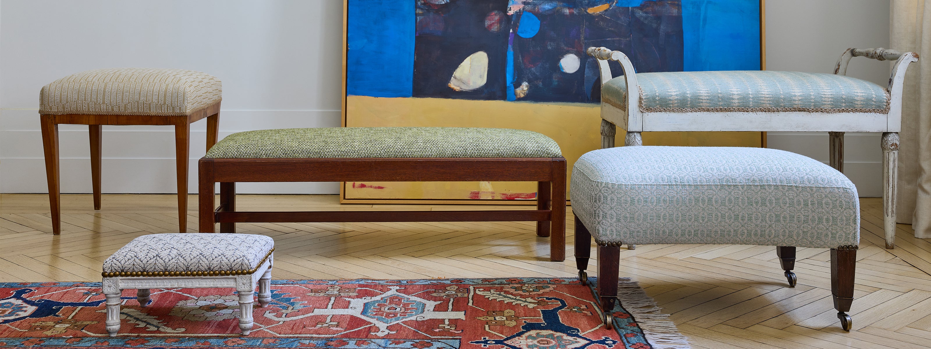A collection of upholstered wooden furniture pieces, including a bench, two stools, and an ottoman, arranged in a styled room setting with a patterned rug and a colorful painting on the wall. Luxury interior design fabrics sold by the metre online and our London showroom.